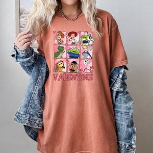 K&ouml;nnte beinhalten: Ein rostfarbenes T-Shirt mit einem Motiv von Toy Story-Charakteren in rosa Herzen. Der Text "VALENTINE" ist unter dem Motiv gedruckt.