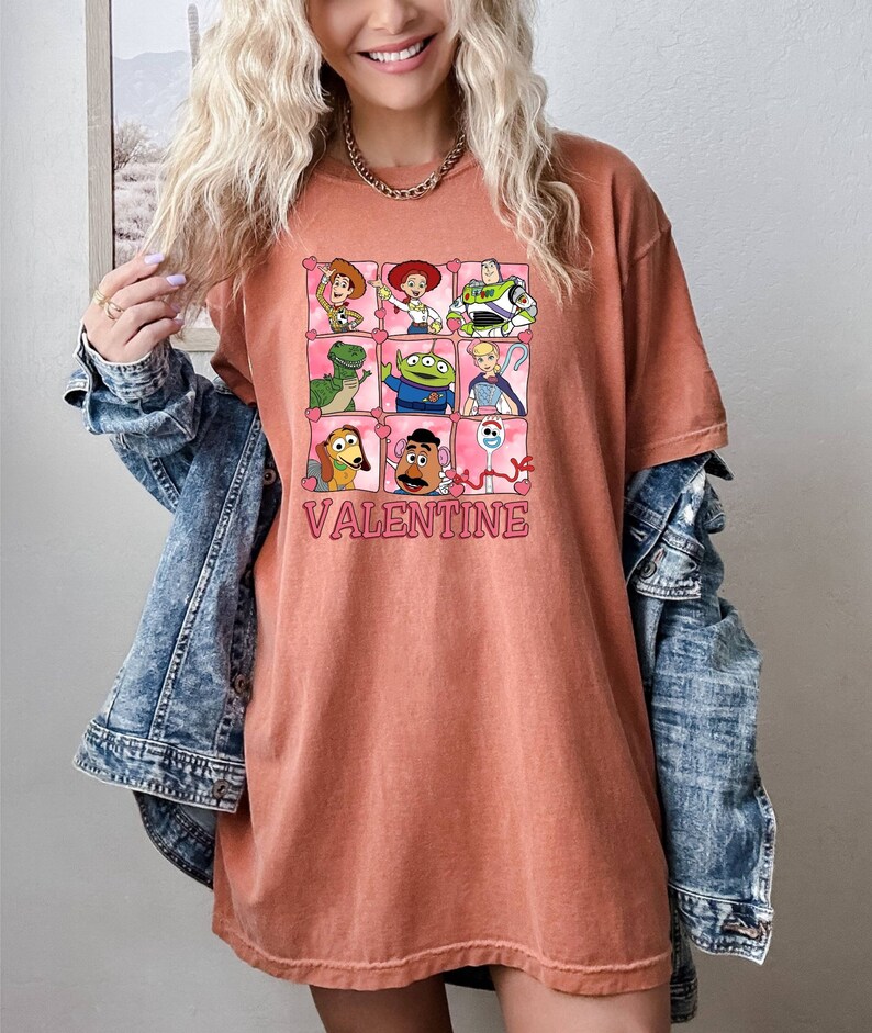 K&ouml;nnte beinhalten: Ein rostfarbenes T-Shirt mit einem Motiv von Toy Story-Charakteren in rosa Herzen. Der Text "VALENTINE" ist unter dem Motiv gedruckt.