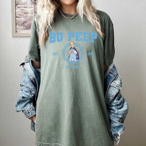 Puede incluir: Camiseta verde de gran tama&ntilde;o con un gr&aacute;fico azul de Bo Peep de Toy Story. El gr&aacute;fico incluye el texto "Bo Peep", "Est. 1995" y "Toy Story".