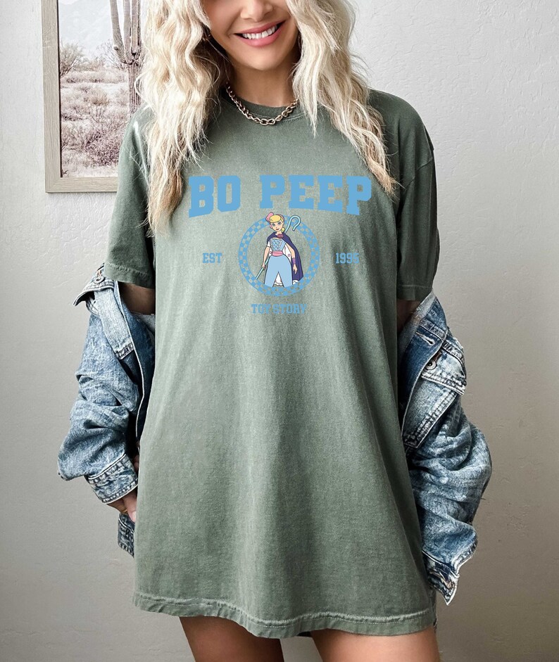 Puede incluir: Camiseta verde de gran tama&ntilde;o con un gr&aacute;fico azul de Bo Peep de Toy Story. El gr&aacute;fico incluye el texto "Bo Peep", "Est. 1995" y "Toy Story".