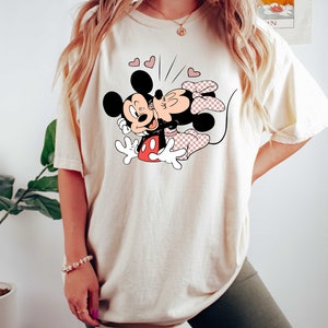 May include: A cream coloured t-shirt featuring a graphic of Mickey Mouse and Minnie Mouse kissing. The graphic is in black, red and pink. There are small pink hearts around the graphic.