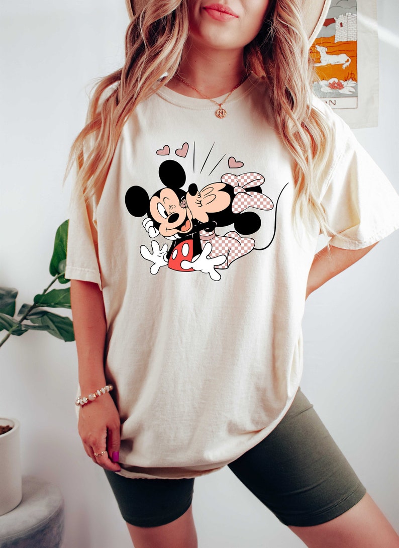 May include: A cream coloured t-shirt featuring a graphic of Mickey Mouse and Minnie Mouse kissing. The graphic is in black, red and pink. There are small pink hearts around the graphic.
