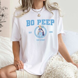 Puede incluir: Camiseta blanca con un estampado gr&aacute;fico azul y blanco de Bo Peep de Toy Story. El gr&aacute;fico incluye el texto "Bo Peep", "Est. 1995", y "Toy Story".