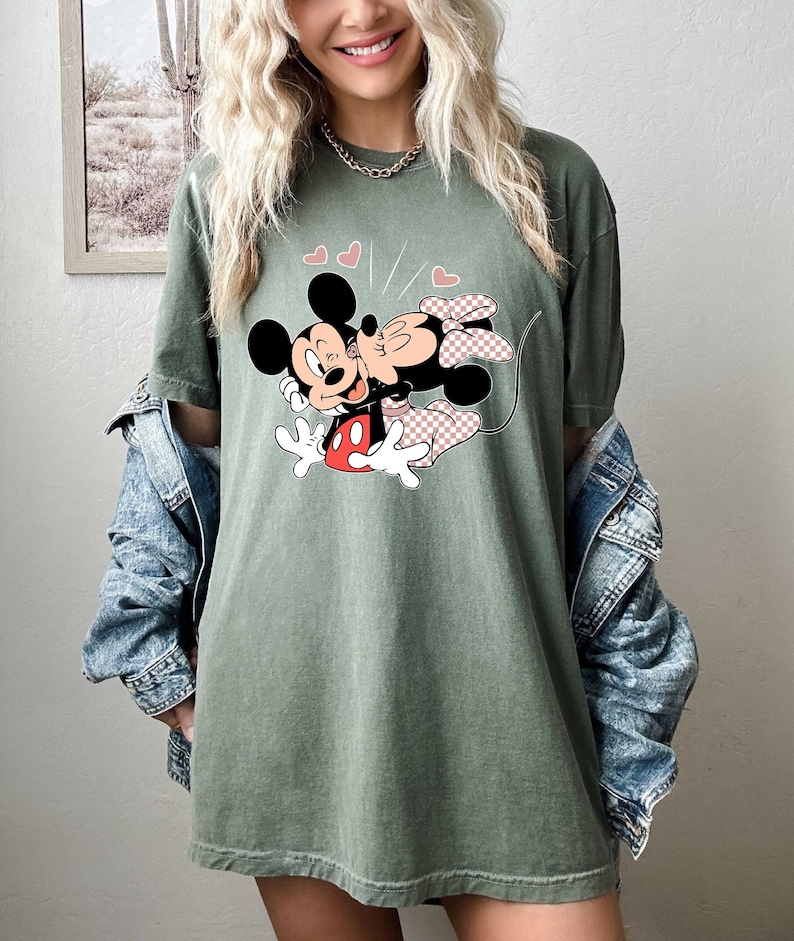 May include: A green t-shirt featuring a graphic of Mickey Mouse and Minnie Mouse kissing. The graphic is in a cartoon style and features red, black and white colours. There are pink hearts above the characters.