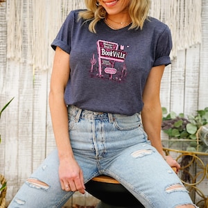 May include: A woman wearing a navy blue t-shirt with a pink graphic of a sign that says "Welcome to Bookville". She is wearing light blue jeans with distressed details.