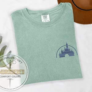 May include: A light green t-shirt with a dark blue embroidered image of a castle. The t-shirt has a crew neck and short sleeves.