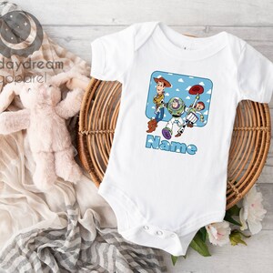 May include: White baby onesie with a cartoon image of Woody, Buzz Lightyear, and Jessie from Toy Story. The image is surrounded by a blue square with white clouds. The text "Name" is printed below the image.