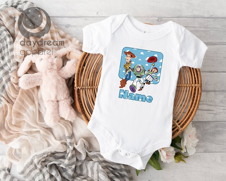May include: White baby onesie with a cartoon image of Woody, Buzz Lightyear, and Jessie from Toy Story. The image is surrounded by a blue square with white clouds. The text "Name" is printed below the image.