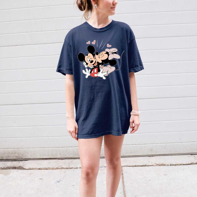 May include: An oversized navy blue t-shirt featuring a graphic of Mickey Mouse and Minnie Mouse kissing. The graphic is in a cartoon style with red, black and white colours. There are three pink hearts above the graphic.