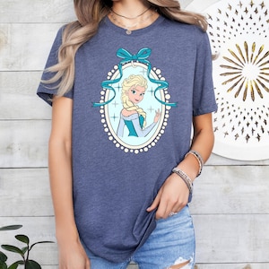 Puede incluir: Una camiseta azul marino con una imagen de dibujos animados de una mujer rubia con un vestido azul y blanco, enmarcada por un &oacute;valo azul claro con un lazo y una cinta. La mujer sonr&iacute;e y saluda. La camiseta es de material suave.