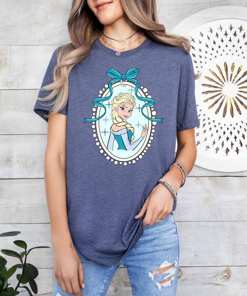 Puede incluir: Una camiseta azul marino con una imagen de dibujos animados de una mujer rubia con un vestido azul y blanco, enmarcada por un &oacute;valo azul claro con un lazo y una cinta. La mujer sonr&iacute;e y saluda. La camiseta es de material suave.