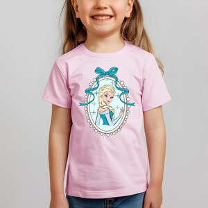 Puede incluir: Camiseta rosa claro con una ilustraci&oacute;n de dibujos animados de una mujer rubia con un vestido azul y blanco, enmarcada por una cinta azul y un borde ovalado. Llevada por una ni&ntilde;a.