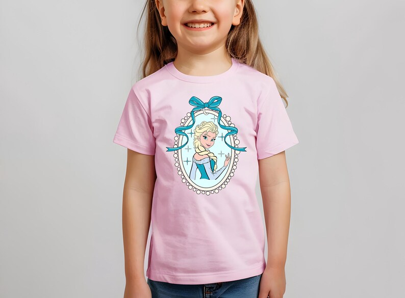 Puede incluir: Camiseta rosa claro con una ilustraci&oacute;n de dibujos animados de una mujer rubia con un vestido azul y blanco, enmarcada por una cinta azul y un borde ovalado. Llevada por una ni&ntilde;a.
