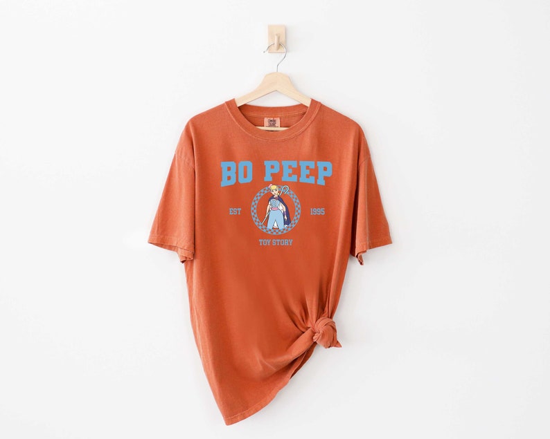Puede incluir: Una camiseta de color &oacute;xido con un gr&aacute;fico azul y blanco de Bo Peep de Toy Story. El gr&aacute;fico incluye el texto "Bo Peep", "Est. 1995", y "Toy Story".