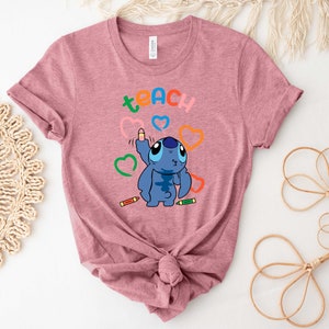 May include: A pink t-shirt featuring a blue Stitch character drawing a heart with a crayon. The word "teach" is written above Stitch in rainbow colours.