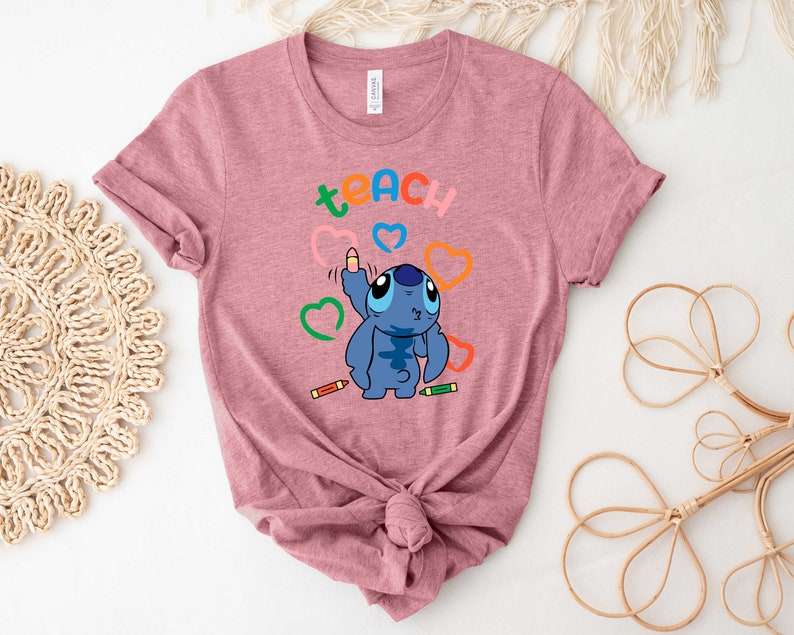 May include: A pink t-shirt featuring a blue Stitch character drawing a heart with a crayon. The word "teach" is written above Stitch in rainbow colours.
