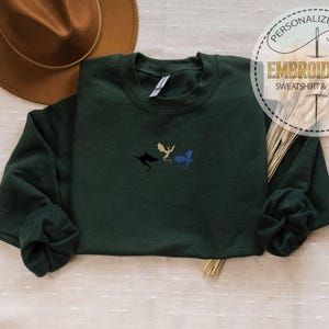 May include: A dark green sweatshirt with embroidered dragon designs in black, gold, and blue. The sweatshirt has long sleeves and a crew neck. The image also includes a brown hat and a decorative circular emblem that reads "PERSONALIZED GIFT EMBROIDERED SWEATSHIRT & HOODIE."
