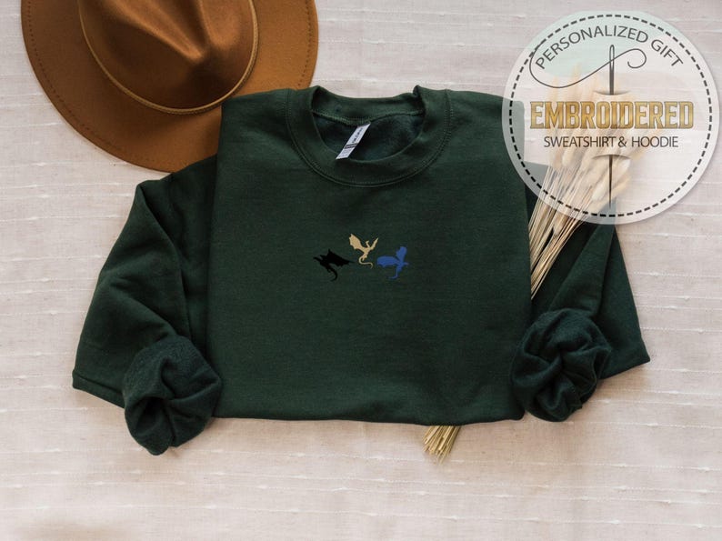 May include: A dark green sweatshirt with embroidered dragon designs in black, gold, and blue. The sweatshirt has long sleeves and a crew neck. The image also includes a brown hat and a decorative circular emblem that reads "PERSONALIZED GIFT EMBROIDERED SWEATSHIRT & HOODIE."