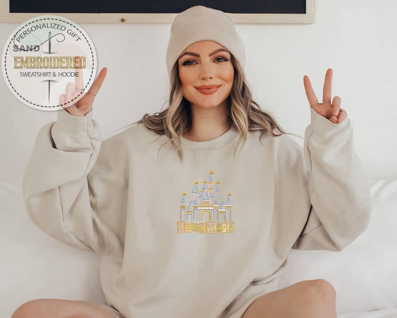 May include: A beige sweatshirt with an embroidered castle design and the words "Disney World" in yellow. The sweatshirt is paired with a matching beanie. The image also includes a circular logo that says "Personalized Gift Sand Embroidered Sweatshirt & Hoodie".