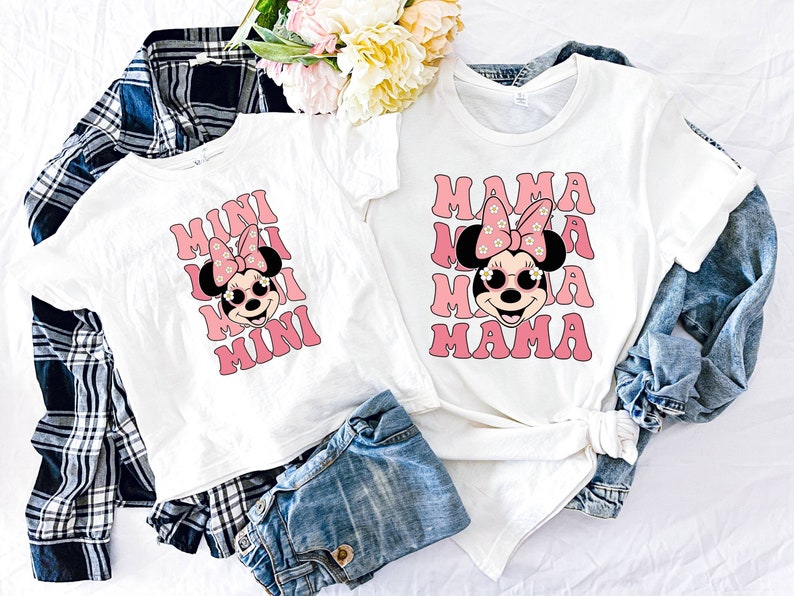 K&ouml;nnte beinhalten: Zwei wei&szlig;e T-Shirts mit einem rosa-wei&szlig;en Minnie Mouse-Design. Das gr&ouml;&szlig;ere Shirt sagt "MAMA" und das kleinere Shirt sagt "MINI".
