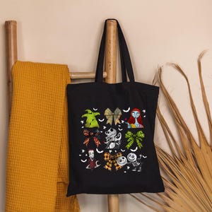 Puede incluir: Bolso tote negro con ilustraciones coloridas de personajes y lazos. El bolso est&aacute; colgado de una escalera de madera junto a una manta de color mostaza. Las ilustraciones incluyen una figura verde, un personaje con cabello rojo y un esqueleto.