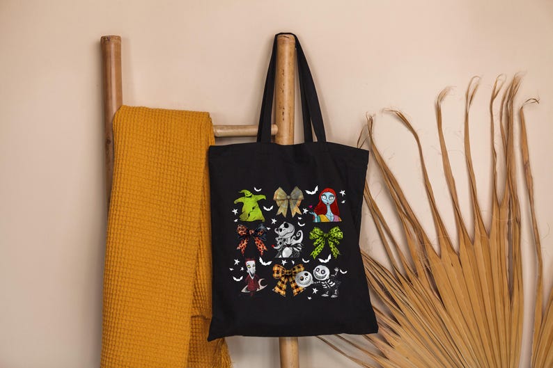 Puede incluir: Bolso tote negro con ilustraciones coloridas de personajes y lazos. El bolso est&aacute; colgado de una escalera de madera junto a una manta de color mostaza. Las ilustraciones incluyen una figura verde, un personaje con cabello rojo y un esqueleto.