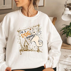 May include: White sweatshirt with a graphic that says "I'M A DOG PERSON". The graphic features a cartoon dog character with a pumpkin nose and a small church. The design is in shades of orange, black, and beige.