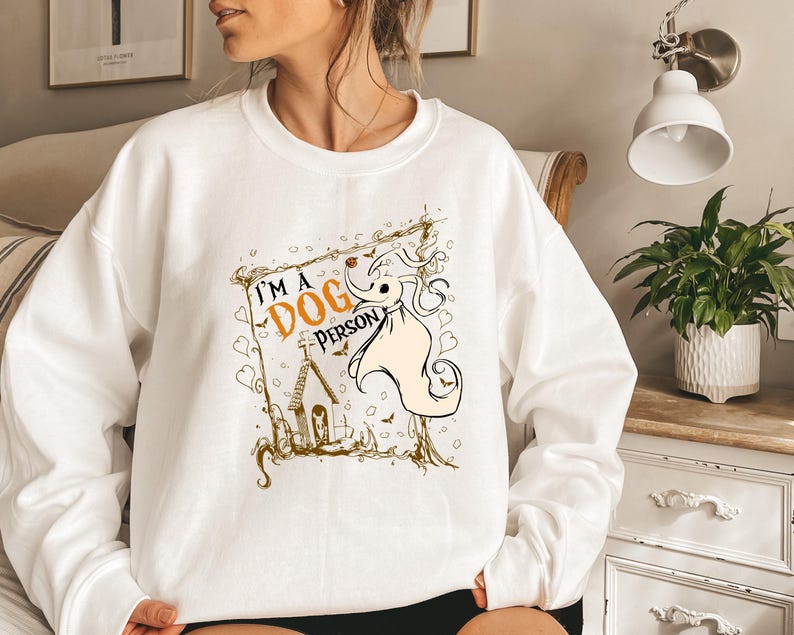 May include: White sweatshirt with a graphic that says "I'M A DOG PERSON". The graphic features a cartoon dog character with a pumpkin nose and a small church. The design is in shades of orange, black, and beige.