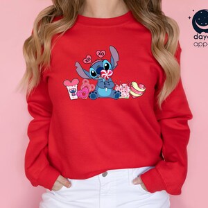 K&ouml;nnte beinhalten: Roter Pullover mit einer blauen Stitch-Figur, die einen Lolli h&auml;lt und von Herzen und S&uuml;&szlig;igkeiten umgeben ist. Der Text "XO" befindet sich auf einem Herz &uuml;ber Stitch's Kopf.