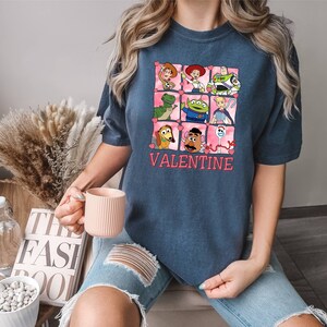 K&ouml;nnte beinhalten: Ein marineblaues T-Shirt mit einer pinkfarbenen und wei&szlig;en Grafik von Toy Story-Charakteren in einem herzf&ouml;rmigen Rahmen. Der Text "VALENTINE" ist unter dem Rahmen gedruckt.