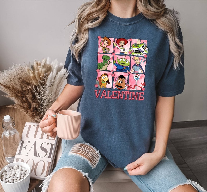 K&ouml;nnte beinhalten: Ein marineblaues T-Shirt mit einer pinkfarbenen und wei&szlig;en Grafik von Toy Story-Charakteren in einem herzf&ouml;rmigen Rahmen. Der Text "VALENTINE" ist unter dem Rahmen gedruckt.