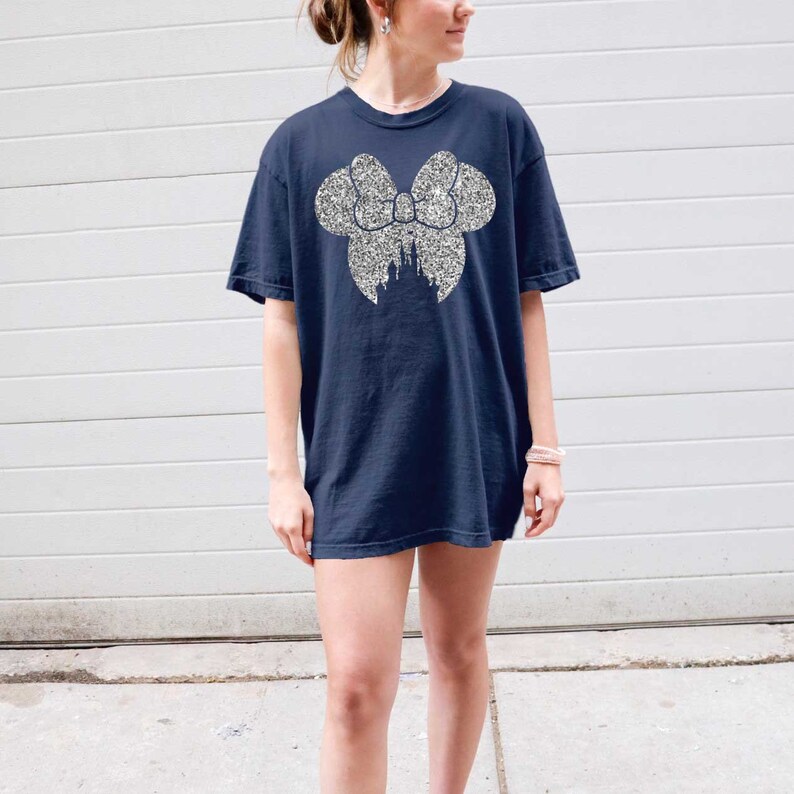 Puede incluir: Una camiseta azul marino con un dise&ntilde;o de purpurina plateada de un lazo de Minnie Mouse con la silueta del castillo de Cenicienta dentro del lazo.