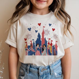 K&ouml;nnte beinhalten: Ein cremefarbenes T-Shirt mit einem patriotischen Design mit Disney-Prinzessinnen. Die Grafik zeigt eine Schlosssilhouette, Feuerwerk und Mickey Mouse-Ballons in Rot und Blau. Die Prinzessinnen sind in rot, wei&szlig; und blauen Outfits gekleidet.