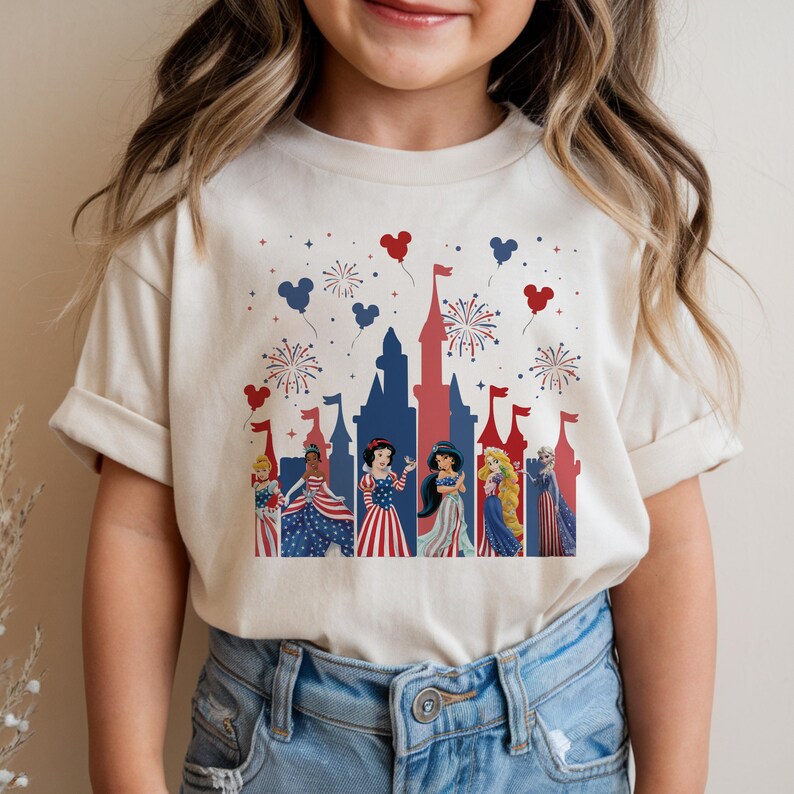 K&ouml;nnte beinhalten: Ein cremefarbenes T-Shirt mit einem patriotischen Design mit Disney-Prinzessinnen. Die Grafik zeigt eine Schlosssilhouette, Feuerwerk und Mickey Mouse-Ballons in Rot und Blau. Die Prinzessinnen sind in rot, wei&szlig; und blauen Outfits gekleidet.