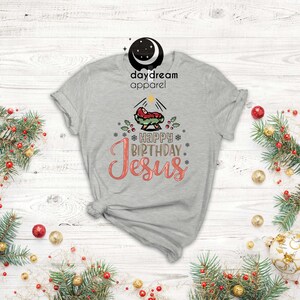 May include: A gray t-shirt with a Christmas nativity scene and the text "Happy Birthday Jesus" in pink and gold lettering.