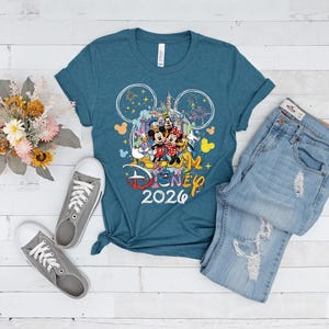 May include: Teal t-shirt with a Disney graphic featuring Mickey Mouse, Minnie Mouse, Donald Duck, and other characters, along with the text "Disney 2026." The shirt is paired with light-wash denim jeans and gray sneakers. A bouquet of dried flowers is also present.