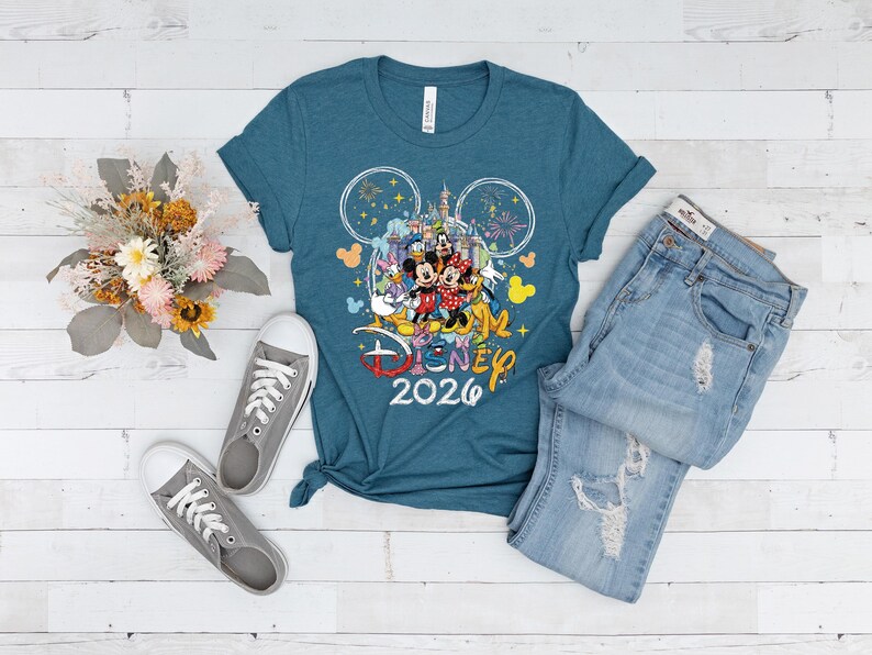 May include: Teal t-shirt with a Disney graphic featuring Mickey Mouse, Minnie Mouse, Donald Duck, and other characters, along with the text "Disney 2026." The shirt is paired with light-wash denim jeans and gray sneakers. A bouquet of dried flowers is also present.