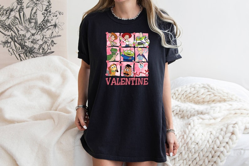 K&ouml;nnte beinhalten: Schwarzes T-Shirt mit einem Aufdruck von Toy Story-Charakteren in rosa Herzen. Der Text "VALENTINE" ist unter dem Aufdruck gedruckt.