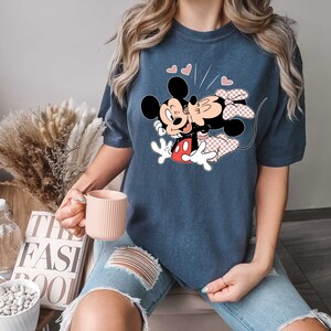 May include: A navy blue t-shirt featuring a graphic of Mickey Mouse and Minnie Mouse kissing. The graphic is in black, red and white. The shirt has a short sleeve and a crew neck.
