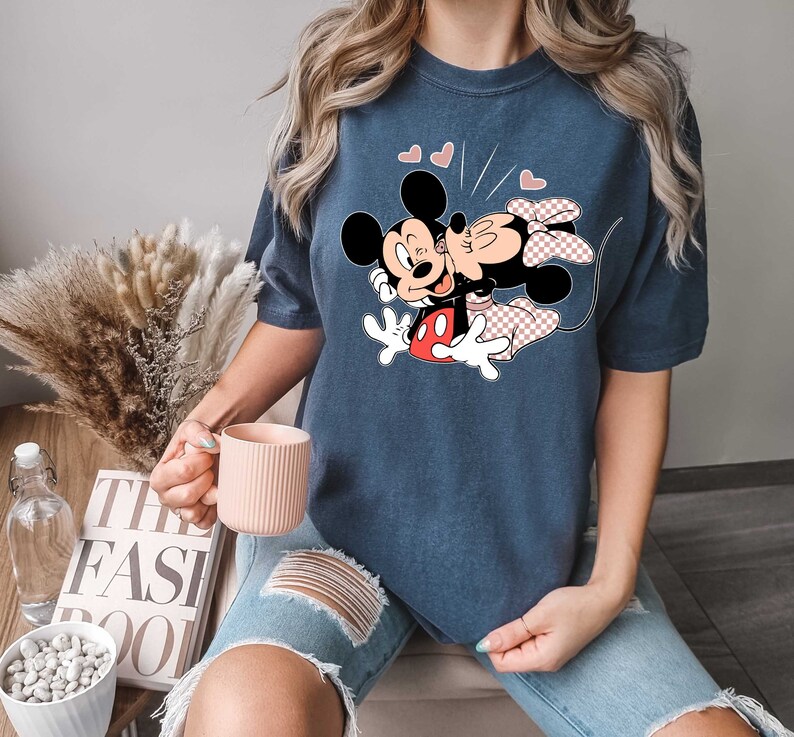May include: A navy blue t-shirt featuring a graphic of Mickey Mouse and Minnie Mouse kissing. The graphic is in black, red and white. The shirt has a short sleeve and a crew neck.