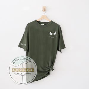 K&ouml;nnte beinhalten: Olivgr&uuml;nes T-Shirt mit einem wei&szlig;en Jack Skellington-Gesicht auf der Brust. Der &Auml;rmel hat den Text "Hor Jack". Das Shirt h&auml;ngt an einem Holzb&uuml;gel vor wei&szlig;em Hintergrund.