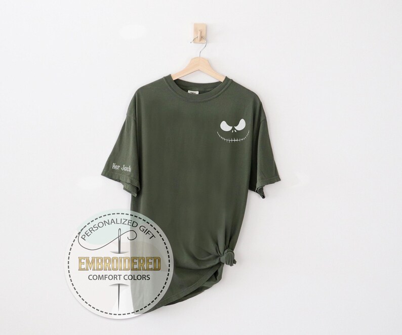 K&ouml;nnte beinhalten: Olivgr&uuml;nes T-Shirt mit einem wei&szlig;en Jack Skellington-Gesicht auf der Brust. Der &Auml;rmel hat den Text "Hor Jack". Das Shirt h&auml;ngt an einem Holzb&uuml;gel vor wei&szlig;em Hintergrund.