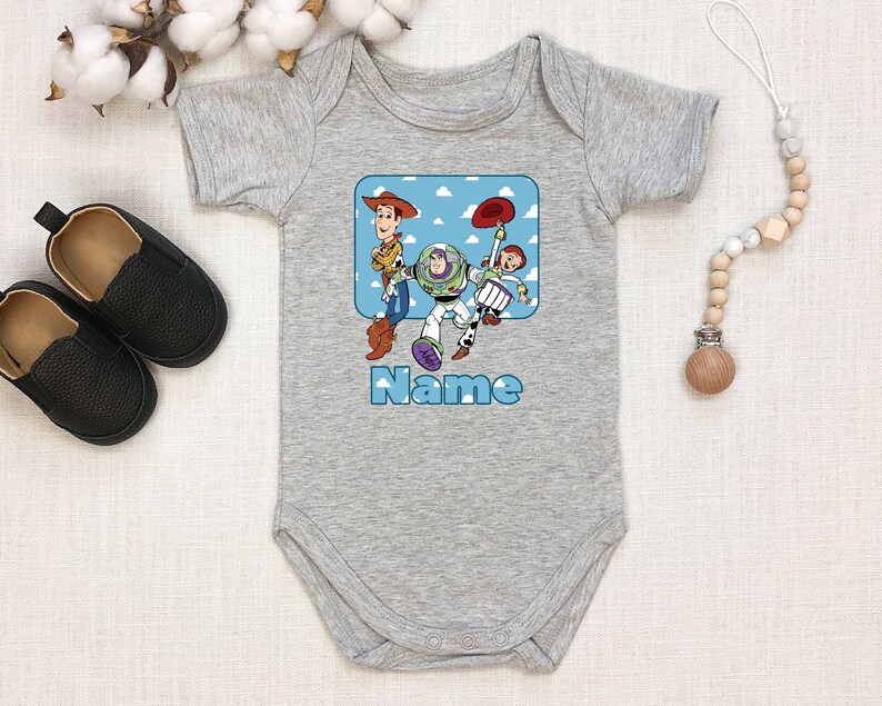 May include: Gray baby onesie with a blue and white cartoon image of Woody, Buzz Lightyear, and Jessie from Toy Story. The text "Name" is printed below the image.