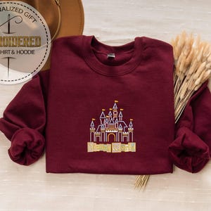 May include: A maroon sweatshirt with a detailed embroidered castle design in gold, purple, and yellow. The text "Disney World" is below the castle. The image also includes a circular logo that says "Personalized Gift" and "Embroidered Sweatshirt & Hoodie".