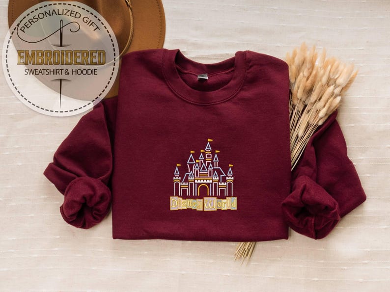 May include: A maroon sweatshirt with a detailed embroidered castle design in gold, purple, and yellow. The text "Disney World" is below the castle. The image also includes a circular logo that says "Personalized Gift" and "Embroidered Sweatshirt & Hoodie".