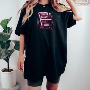 May include: A black oversized t-shirt with a pink graphic of a neon sign that reads "Welcome Read On to Bookville".