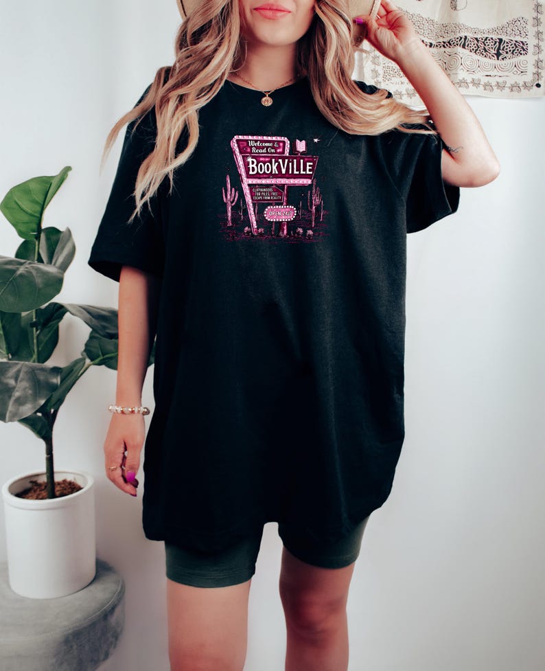 May include: A black oversized t-shirt with a pink graphic of a neon sign that reads "Welcome Read On to Bookville".