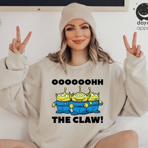 K&ouml;nnte beinhalten: Ein hellbeiger Pullover mit einem Aufdruck von drei gr&uuml;nen Au&szlig;erirdischen, die jeweils drei Augen haben. Die Au&szlig;erirdischen tragen blaue Overalls und stehen in einer Reihe. Der Text "Oooooohhh The Claw!" ist unter den Au&szlig;erirdischen aufgedruckt.