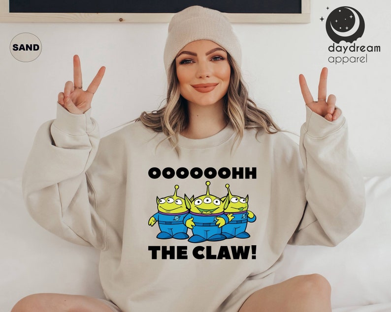 K&ouml;nnte beinhalten: Ein hellbeiger Pullover mit einem Aufdruck von drei gr&uuml;nen Au&szlig;erirdischen, die jeweils drei Augen haben. Die Au&szlig;erirdischen tragen blaue Overalls und stehen in einer Reihe. Der Text "Oooooohhh The Claw!" ist unter den Au&szlig;erirdischen aufgedruckt.