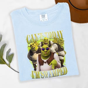 May include: Pale blue t-shirt with a graphic of Shrek and the text "CAN'T TODAY I'M SWAMPED" in gold lettering. The image shows Shrek wearing sunglasses with other Shrek characters. The shirt is made of a soft material.